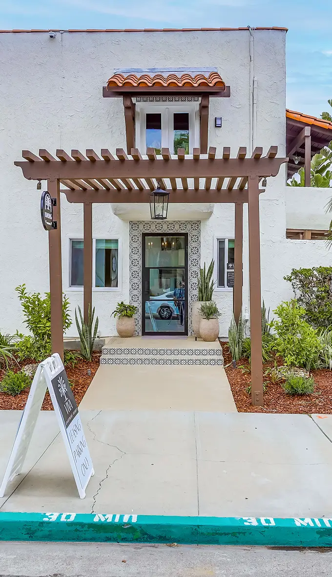 A Spanish-style building with a pergola entrance, decorative tiles, and lush greenery. A sign near the sidewalk reads 'Thank you for visiting.' Let me know if you need refinements!