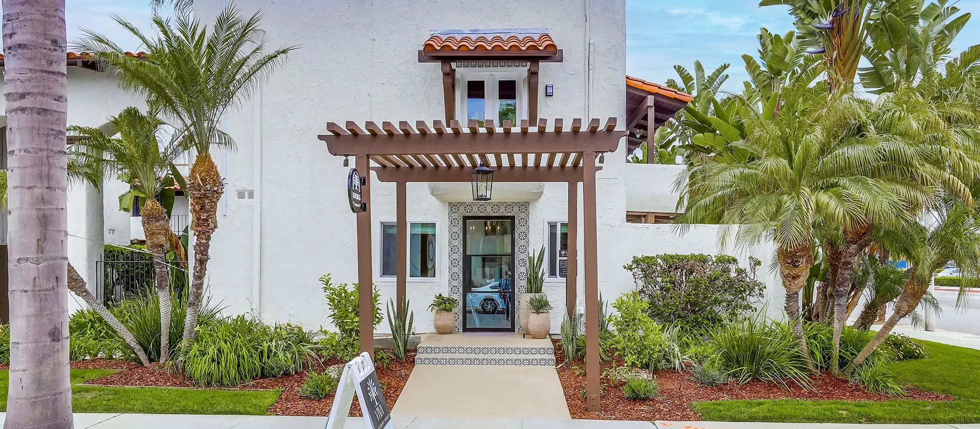 A Spanish-style building with a pergola entrance, decorative tiles, and lush greenery. A sign near the sidewalk reads. 'Thank you for visiting.' Let me know if you need refinements!